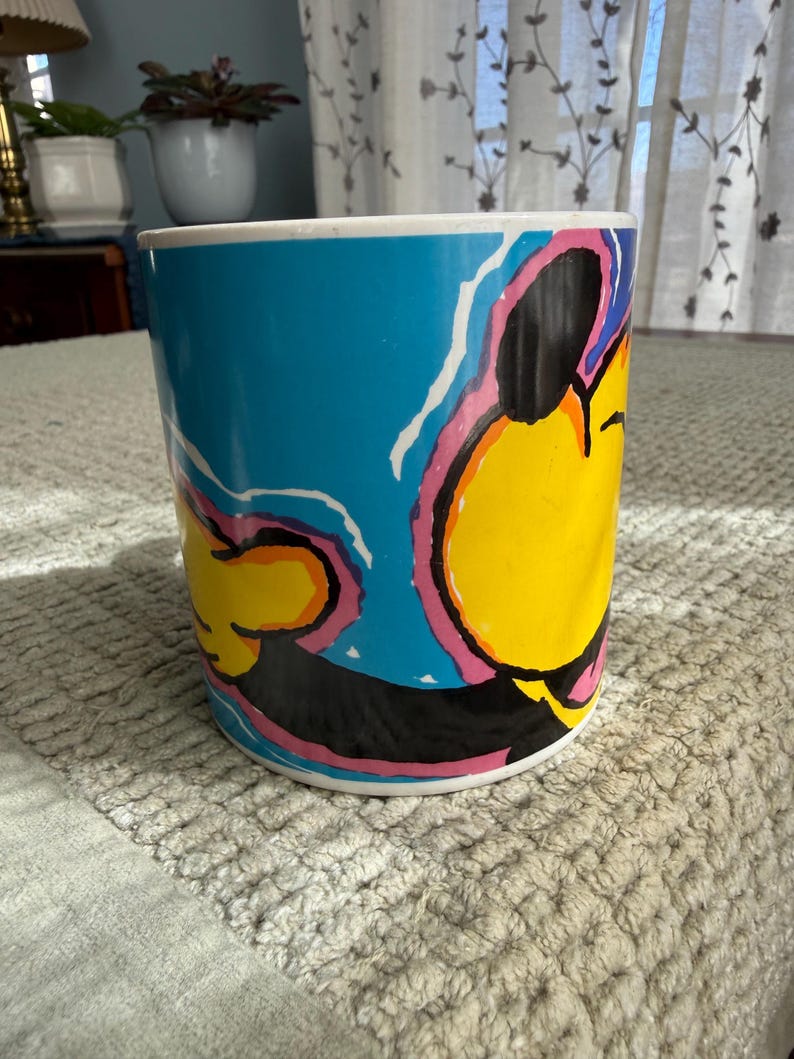 Peut inclure: Une tasse cylindrique en c&eacute;ramique avec un design pop art vibrant. La tasse pr&eacute;sente un fond bleu vif avec un motif de personnage de dessin anim&eacute; jaune, noir, rose et blanc. La tasse a un bord et une base blancs.