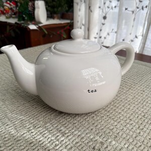May include: A white ceramic teapot with a rounded body, a curved spout, and a loop handle. The lid has a small knob. The word "tea" is printed in black on the front of the teapot. The teapot is on a textured, light-coloured surface.