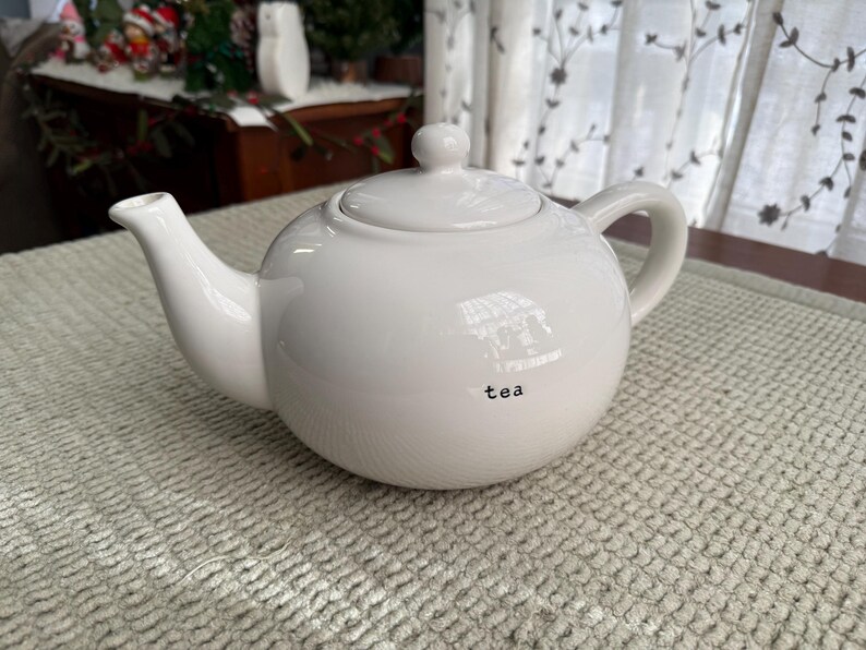 May include: A white ceramic teapot with a rounded body, a curved spout, and a loop handle. The lid has a small knob. The word "tea" is printed in black on the front of the teapot. The teapot is on a textured, light-coloured surface.