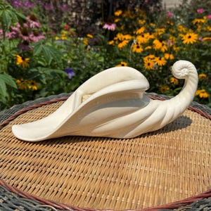 May include: A cream-colored ceramic cornucopia vessel featuring a smooth, glossy finish and a spiraled, horn-shaped design that curves upward at the tail. The object has a wide, open mouth and a tapered, curled end, showcasing a sculptural form.