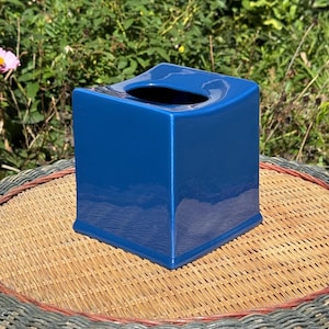 May include: A square, glossy, deep blue tissue box cover with a smooth, reflective finish. The object features a centred, rounded rectangular opening at the top for dispensing tissues, with clean, straight edges and a slightly flared base.