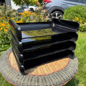 May include: A stack of four black plastic document trays with a glossy finish. Each tray features a rectangular shape with curved front edges and open sides, designed for organizing papers and files in a tiered, vertical arrangement.
