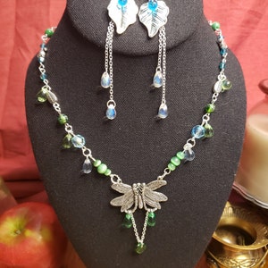 以下が含まれることがあります： トンボのペンダントと緑と青のガラスビーズが付いたシルバーネックレス。葉のデザインと青いガラスのアクセントが付いたシルバーイヤリングがセットになっています。