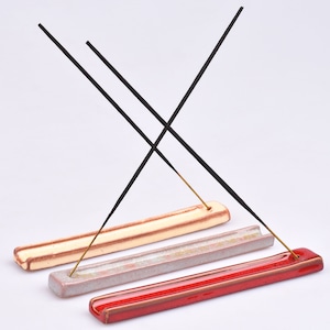 Könnte beinhalten: Drei Keramik-Räucherstäbchenhalter in Rot, Weiß und Beige mit brennenden schwarzen Räucherstäbchen. Die Halter sind rechteckig und leicht gebogen.