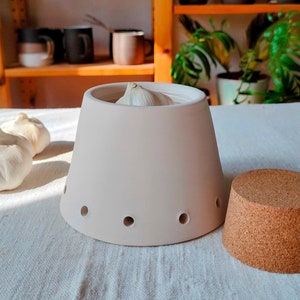 May include: A white ceramic garlic keeper with a cork lid and a single clove of garlic inside. The keeper has holes for ventilation and is sitting on a white linen tablecloth.