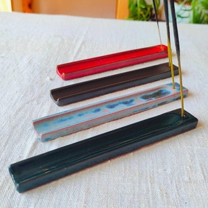 Könnte beinhalten: Vier Keramik-Räucherstäbchenhalter in verschiedenen Farben, darunter Schwarz, Blau, Rot und Grün. Die Halter sind in einer Reihe auf einer weißen Oberfläche angeordnet. Zwei Räucherstäbchen stehen aufrecht im Halter.