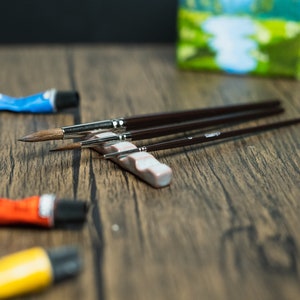 Light blue paint brush holder, brushes, paint and small painting on wooden table.