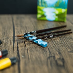 Light blue paint brush holder, brushes, paint and small painting on wooden table.