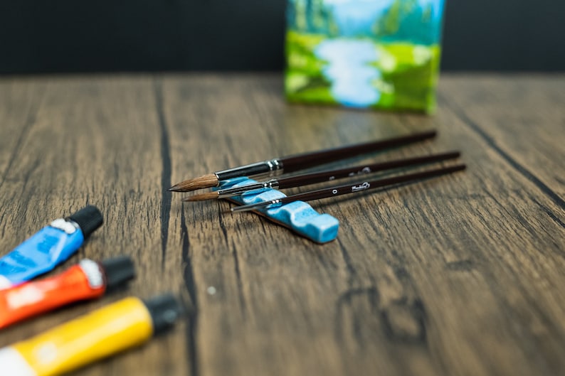 Light blue paint brush holder, brushes, paint and small painting on wooden table.