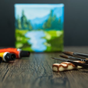 Brown paint brush holder, brushes, paint and small painting on wooden table.