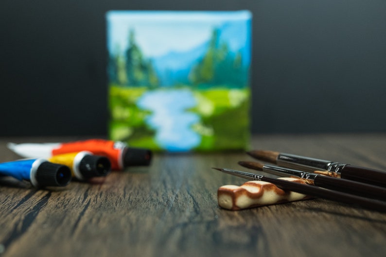 Brown paint brush holder, brushes, paint and small painting on wooden table.