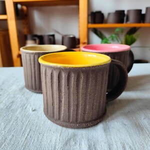 Puede incluir: Tres tazas de cerámica con exteriores texturizados marrones e interiores esmaltados de colores. Las tazas tienen un esmalte amarillo, rosa y beige en el interior. Las tazas están sobre una superficie texturizada blanca.