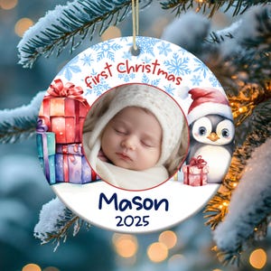 Op de afbeelding: Een witte ronde kerstbal met een blauw sneeuwvlokkenpatroon. Het heeft een foto van een baby in een witte muts, omringd door cadeaus en een pinguïn met een kerstmanmuts. De tekst "first Christmas" is in rood en blauw geschreven, en de naam van de baby "Mason" en het jaar "2025" zijn in blauw geschreven.