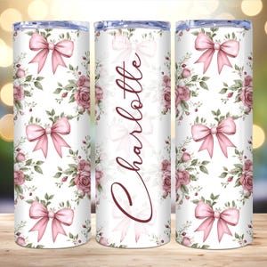 May include: A white tumbler with a floral pattern and pink bows. The tumbler has the name "Charlotte" written on it in a burgundy font.