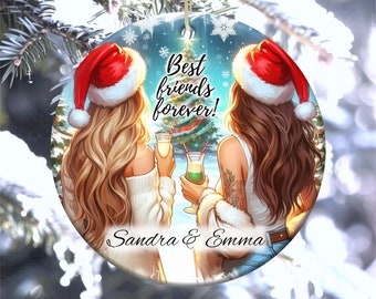 May include: Round Christmas ornament featuring a winter scene with two women wearing Santa hats and holding drinks. The text "Best friends forever!" is written in white on a blue background. The names "Sandra & Emma" are written at the bottom of the ornament.