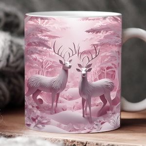 Puede incluir: Una taza de cer&aacute;mica blanca con un dise&ntilde;o de papel recortado rosa de dos ciervos de pie en un bosque nevado. Los ciervos tienen cuernos y se miran el uno al otro.