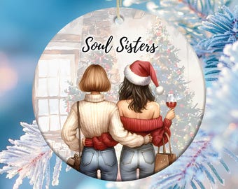 May include: A Christmas ornament featuring two women standing in front of a Christmas tree. The women are wearing jeans and sweaters. One woman is wearing a Santa hat. The ornament says "Soul Sisters" at the top.