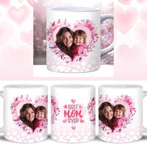 Op de afbeelding: Witte keramische mok met een roze hartontwerp met een foto van een moeder en dochter. Het hart is omgeven door roze bloemen en bladeren. De tekst "Best Mom Ever" is in roze op de mok gedrukt.