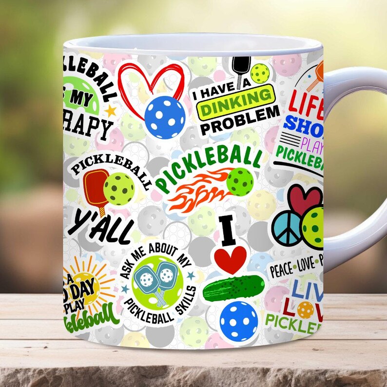 Puede incluir: Taza de caf&eacute; de cer&aacute;mica blanca con un dise&ntilde;o colorido que presenta raquetas de pickleball, pelotas y frases como "Tengo un problema de dinking" y "La vida es corta, juega pickleball".