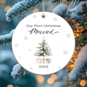 May include: White round Christmas ornament with a watercolor illustration of a Christmas tree with presents underneath. The ornament has the text "Our First Christmas Married 2025" on it.