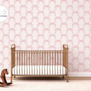 May include: A wooden cot with a white mattress sits in front of a light pink wallpaper featuring a repeating pattern of small pink bows. A small wooden rocking horse is positioned to the left of the cot. The logo Rachel Lin Design is visible.