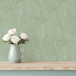 Puede incluir: Papel pintado verde claro con un patrón de cactus blanco. Una jarra azul claro con rosas blancas se encuentra sobre una superficie de madera. El diseño del papel pintado presenta varias formas de cactus.