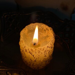 May include: A lit, textured candle with a warm, yellow glow. The candle has a rough, organic appearance and sits on a decorative, dark-toned tray. The flame is bright and centered, creating a cozy ambiance.