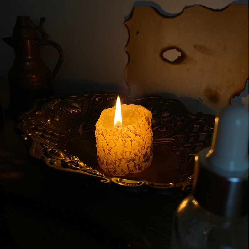 May include: A lit, textured, yellow candle sits on an ornate, antique-style metal tray. The candle's flame glows brightly, casting a warm light. A dark, vintage-style setting includes a decorative pitcher and a piece of aged paper.