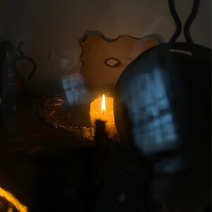 May include: A lit candle with a bright yellow flame, casting a warm glow in a dimly lit setting. The candle is positioned near a decorative object and a metal pitcher. The scene evokes a sense of tranquility and ambiance.