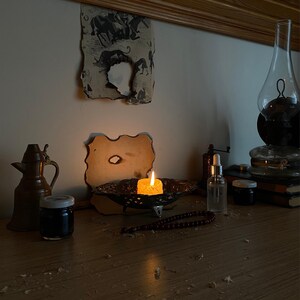May include: A dimly lit scene featuring a burning candle in a decorative bowl, a vintage oil lamp, and a small jar. A string of beads and a small bottle with a dropper are also visible. The setting evokes a sense of history and tranquility.