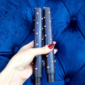 May include: Two dark brown decorative candle holders with iridescent gems. The candle holders are cylindrical and are held against a blue velvet background.