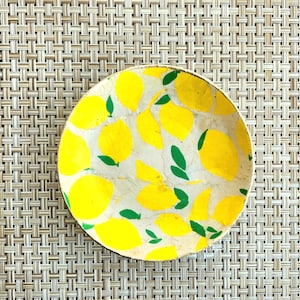以下が含まれることがあります： 白い背景に黄色のレモンと緑の葉の模様が描かれた、丸い装飾的なボウル。ボウルは織り込まれたベージュの表面に置かれています。ボウルのデザインは、夏の柑橘系のテーマを連想させます。
