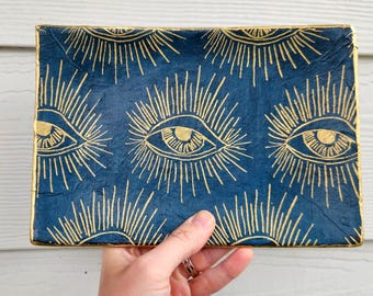 May include: A blue rectangular tray with a gold eye pattern. The tray is made of a textured material and has a slightly raised edge.