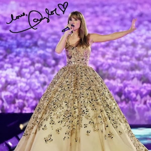 May include: A woman in a gold sequined gown with a long skirt, singing into a microphone on stage. The dress is adorned with intricate gold and black embellishments. The background is a blurred purple hue, with the signature "love, Taylor" visible.