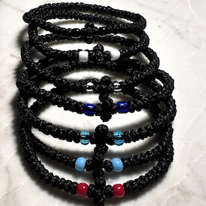 Könnte beinhalten: Eine Reihe schwarzer geknoteter Armbänder mit kreuzförmigen Akzenten. Einige Armbänder haben farbige Perlen in Blau, Rot und Weiß. Die Armbänder liegen auf einer hellen Oberfläche.