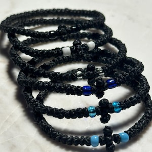 以下が含まれることがあります： 黒色の編み込みブレスレットの束。十字型のアクセントが付いています。一部のブレスレットには、白、青、透明の小さなビーズが付いています。明るい色の表面に表示されています。