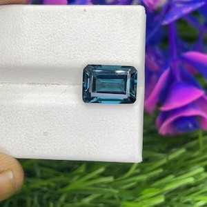 Könnte beinhalten: Ein rechteckiger, teal-farbener Edelstein mit Stufenschliff, präsentiert auf einer weißen Oberfläche. Der Edelstein hat eine tiefe, gesättigte Farbe und eine reflektierende Oberfläche. Der Hintergrund zeigt grünes und violettes Laub.