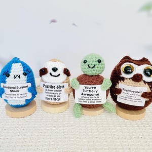 May include: Four crocheted animal plushies holding signs with motivational messages. A blue shark says "Emotional Support Shark", a brown sloth says "Positive Sloth", a green turtle says "You're Turtle-y Awesome", and a brown owl says "Positive Owl".