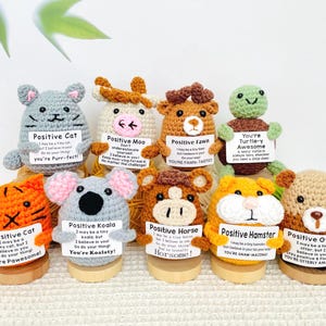 May include: A collection of handmade crochet animal figures, including a cat, cow, deer, turtle, tiger, koala, horse, hamster, and otter. Each figure holds a sign with an inspirational message. The figures are displayed on small wooden bases.