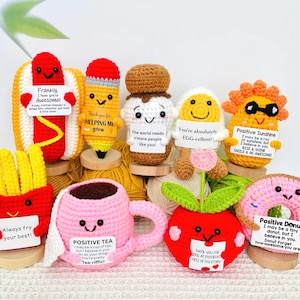 May include: A collection of handmade crochet food items, including a hot dog, pencil, ice cream, egg, sunshine, french fries, tea cup, apple, and donut. Each item has a smiling face and a small sign with a positive message.