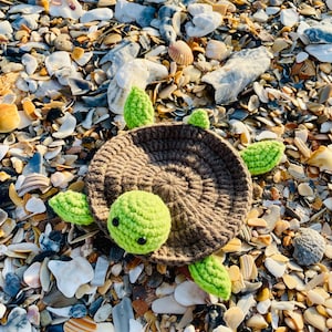 May include: A handmade crochet turtle with a brown shell and bright green head and flippers. The turtle is lying on a beach covered in seashells and pebbles. The turtle's shell is a circular shape, and the head and flippers are rounded.