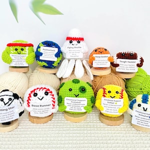 May include: A collection of handmade crochet sports-themed characters, including a tennis ball, baseball, football, and others. Each character has a unique design and a small sign with inspirational text. The characters are displayed with yarn balls.
