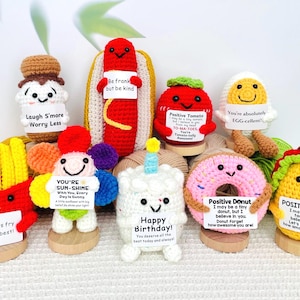 May include: A collection of colorful, crocheted food-themed characters, including a hot dog, fries, a tomato, a donut, and a taco. Each character holds a sign with an inspirational message. The characters are displayed on wooden bases.