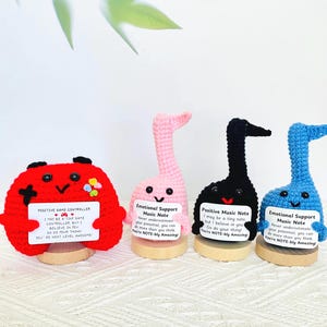 May include: Crocheted figures of a red game controller and three musical notes in pink, black, and blue. Each figure holds a small white sign with inspirational text. The figures are on wooden bases.