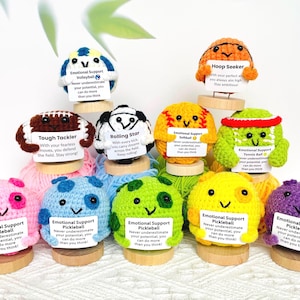 May include: A collection of colorful, crocheted sports-themed characters, each with a unique design and a small sign. The characters include a football, baseball, and volleyball, with encouraging messages. The characters are displayed on wooden bases.