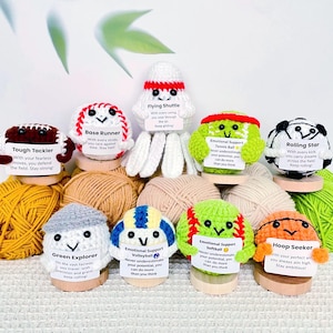 May include: A collection of handmade crochet sports-themed characters, including a football, baseball, and tennis ball, each with a unique design and encouraging message. The characters are displayed on wooden stands, surrounded by yarn.