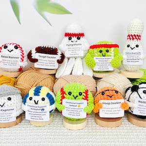 May include: A collection of handmade crochet sports-themed characters, including a baseball, football, tennis ball, bowling pin, and others. Each character has a unique design and a small sign with an inspirational message. The characters are displayed on wooden bases.