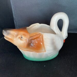 May include: A white ceramic swan with a brown and white fox head inside. The swan has a green base and is a decorative piece.