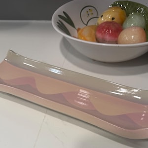 May include: A rectangular ceramic serving tray with a wavy design in shades of pink, peach, and beige. The tray has a glossy finish and gently curved edges. In the background, a white bowl holds colorful, round food items.