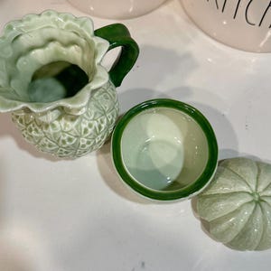Może przedstawiać: Zestaw trzech zielonych ceramicznych artykuł&oacute;w kuchennych. Artykuły mają kształt ananasa i mają fakturowaną powierzchnię. Zestaw zawiera dzbanek, miskę i pokrywkę.